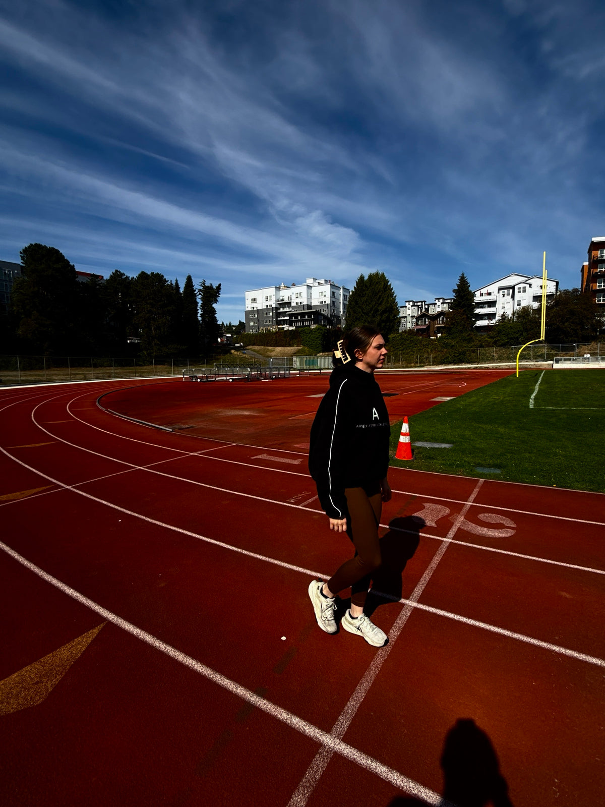 The Apex Hoodie in black worn walking on a red running track under a clear sky, with apartment buildings and evergreens behind.
