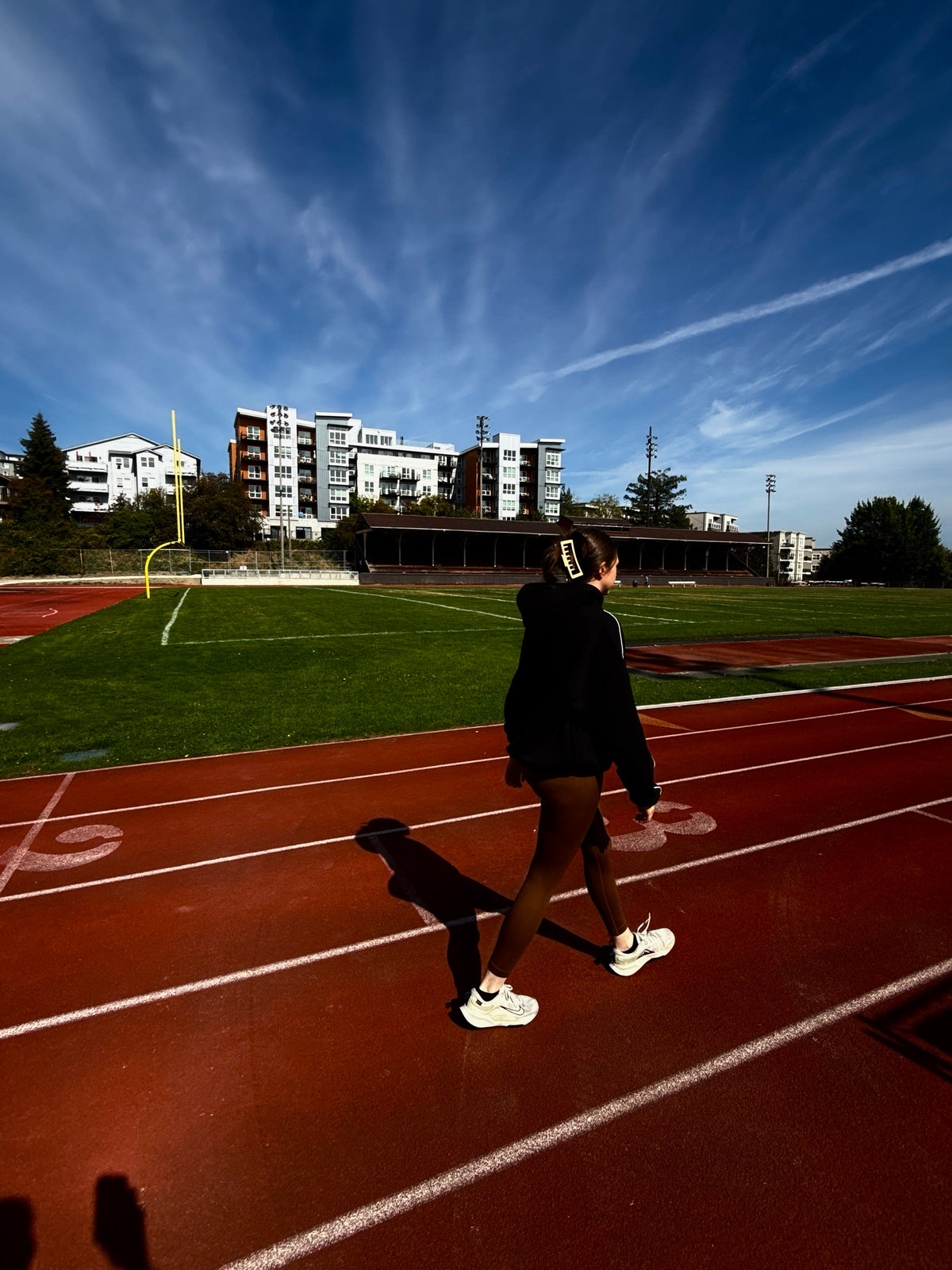 Wide shot of a running track and football field in a Pacific Northwest city, with a person walking the lanes in the black Apex Hoodie.